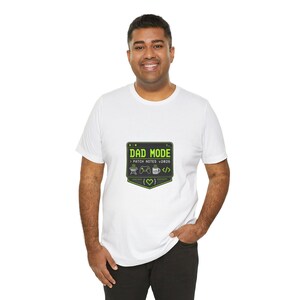 May include: A white short-sleeved t-shirt featuring a dark green graphic shaped like a computer window. The text reads DAD MODE and PATCH NOTES v2026, accompanied by icons of a grill, a game controller, a coffee mug, and code brackets with a heart.