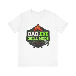 May include: A white short-sleeved t-shirt featuring a graphic of a black charcoal grill with orange flames. The text reads DAD.EXE GRILL MODE ACTIVATED in white and green lettering, accompanied by a computer mouse cursor icon shaped like a spatula.