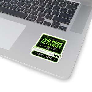 May include: A rectangular sticker with a black background and bright green pixelated text that reads DAD MODE ACTIVATED, featuring a small grill and spatula icon, with a secondary smaller bar below labelled PATCH NOTES in white text.