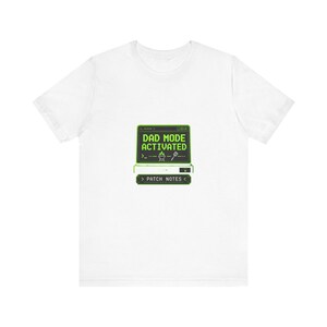 Könnte beinhalten: Ein weißes kurzärmeliges T-Shirt mit einer Grafik eines Computerbildschirms, auf dem in grüner Schrift DAD MODE ACTIVATED steht, dazu ein kleines Grill- und Pfannenwender-Symbol sowie ein grüner Button mit dem Text PATCH NOTES.