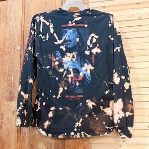 Peut inclure: Un t-shirt noir à manches longues avec un motif délavé à l'eau de Javel. Le dos présente un graphique d'une personne avec des accents bleus et rouges, le texte "THE HELL WE CREATE" en haut et "FIT FOR A KING" en bas, avec du texte sur la manche.