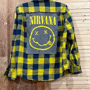 May include: A long-sleeve button-down shirt featuring a yellow and navy blue plaid pattern. The back of the shirt displays a large, rectangular gray fabric patch with the yellow text NIRVANA and a yellow smiley face graphic with X eyes.
