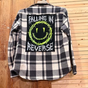 Può includere: Una camicia a maniche lunghe a quadri bianchi e neri con una grande toppa rettangolare sul retro. La toppa nera presenta un grafico di una faccina sorridente in filo spinato verde brillante e la scritta "FALLING IN REVERSE" in bianco.