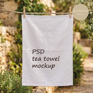 Könnte beinhalten: Ein weißes, rechteckiges Geschirrtuch, das mit einer hölzernen Wäscheklammer an einer Wäscheleine hängt. Das Tuch zeigt den schwarzen aufgedruckten Text "PSD tea towel mockup" mittig auf der Oberfläche. Der Stoff wirkt schlicht und glatt.