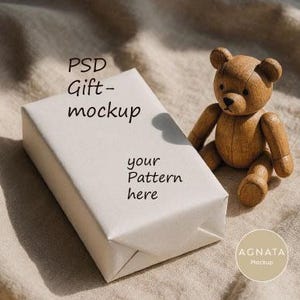 Könnte beinhalten: Eine rechteckige Geschenkbox, eingewickelt in schlichtes cremefarbenes Papier mit dem schwarzen Text PSD Gift-mockup und your Pattern here, begleitet von einer kleinen braunen Teddybär-Figur aus Holz. Ein kreisförmiges Logo mit dem Text AGNATA Mockup ist sichtbar.