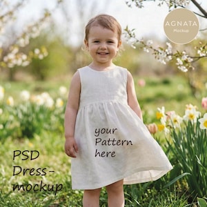 Könnte beinhalten: Ein junges Kind trägt ein weißes ärmelloses Kleid mit ausgestelltem Rock und steht in einem Blumenfeld. Das Kleid hat den Text "your Pattern here" auf dem Rock. Das Bild enthält auch den Text "PSD Dress-mockup" und ein Logo mit der Aufschrift "AGNATA Mockup".