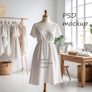 May include: A white dress with short sleeves and a square neckline is displayed on a mannequin. The dress has a fitted waist and a flared skirt. The text "your pattern here" is printed on the skirt. Other white dresses hang on a wooden rack in the background.