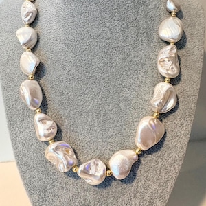May include: A necklace featuring large, irregular white baroque pearls alternating with small, polished gold-tone spherical beads. The pearls have a lustrous, textured surface and are strung together in a single strand to form a continuous circular design.
