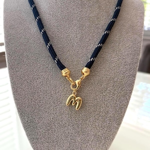 May include: A navy blue rope necklace with silver accents and a gold-colored clasp. The necklace features a gold-colored pendant in the shape of the letter "M". The necklace is displayed on a gray mannequin.