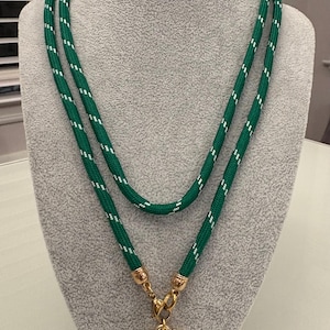 May include: A green rope necklace with white accents and a gold heart pendant. The necklace is displayed on a gray mannequin. The heart pendant is a shiny gold color.