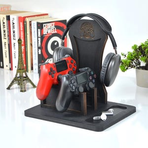 Könnte beinhalten: Schwarzer Holz-Gaming-Headset- und Controller-Ständer. Der Ständer hält ein schwarzes Headset, zwei Gamecontroller (einer rot, einer schwarz) und ein Paar weiße Ohrhörer. Eine kleine Pflanze und eine Astronautenfigur sind ebenfalls zu sehen.