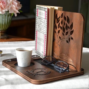 Può includere: Un organizer da scrivania in legno marrone scuro con un fermalibri con un design ad albero. Contiene un libro, una tazza di tè, una penna e occhiali. L'organizer ha un'incisione personalizzata.