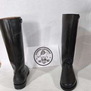 K&ouml;nnte beinhalten: Zwei schwarze Lederreitstiefel. Die Stiefel sind hoch, reichen bis zum Knie, mit glatter Oberfl&auml;che. Eine wei&szlig;e Karte mit einem Logo mit der Aufschrift "COUNTRY LASTSHOES" befindet sich zwischen den Stiefeln.