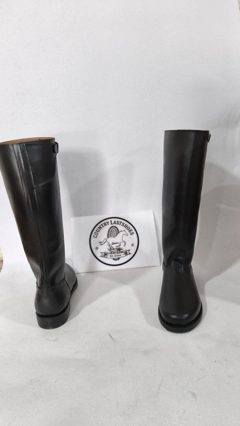 K&ouml;nnte beinhalten: Zwei schwarze Lederreitstiefel. Die Stiefel sind hoch, reichen bis zum Knie, mit glatter Oberfl&auml;che. Eine wei&szlig;e Karte mit einem Logo mit der Aufschrift "COUNTRY LASTSHOES" befindet sich zwischen den Stiefeln.