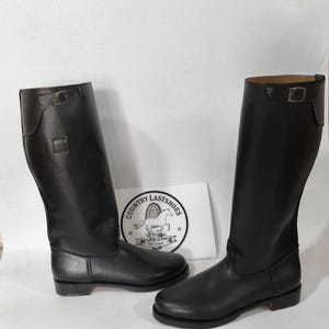 K&ouml;nnte beinhalten: Ein Paar schwarze Lederreitstiefel. Die Stiefel sind hoch, mit glatter Oberfl&auml;che und Schnallendetail am oberen Rand. Sie werden auf einer wei&szlig;en Oberfl&auml;che pr&auml;sentiert.