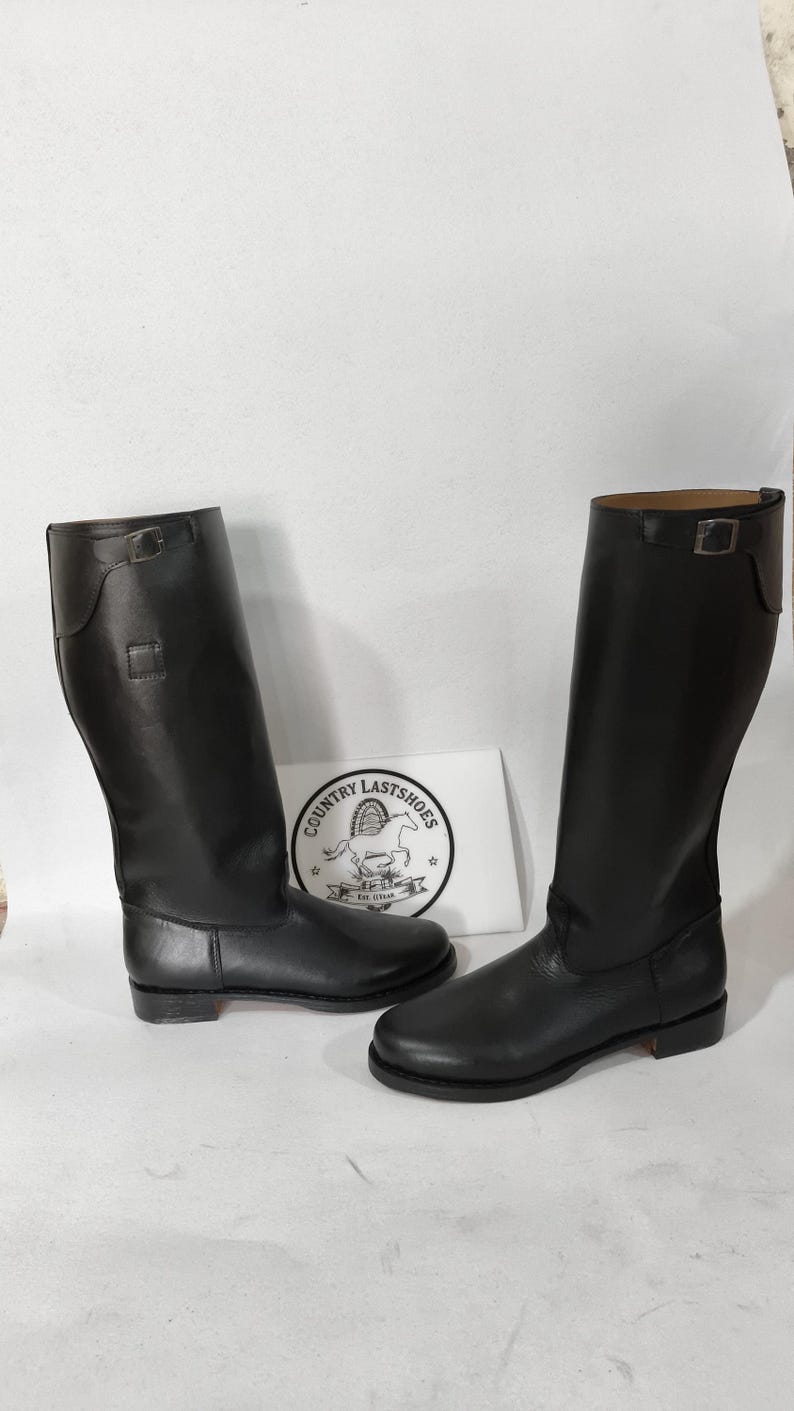 K&ouml;nnte beinhalten: Ein Paar schwarze Lederreitstiefel. Die Stiefel sind hoch, mit glatter Oberfl&auml;che und Schnallendetail am oberen Rand. Sie werden auf einer wei&szlig;en Oberfl&auml;che pr&auml;sentiert.