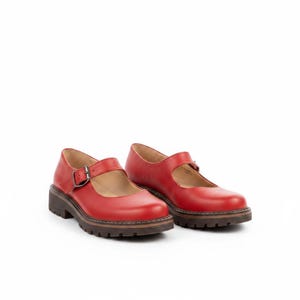 May include: A pair of red leather Mary Jane shoes featuring a rounded toe, a single strap across the instep with a metal buckle closure, and a thick, dark brown lugged rubber sole with visible stitching along the welt of the shoe base.
