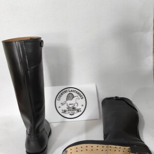 K&ouml;nnte beinhalten: Schwarze Lederreitstiefel, einer aufrecht und einer liegend, mit heller Sohle. Die Stiefel haben einen hohen Schaft und eine runde Spitze. Ein wei&szlig;es Schild mit dem Text "COUNTRY LASTSHOES" ist sichtbar.