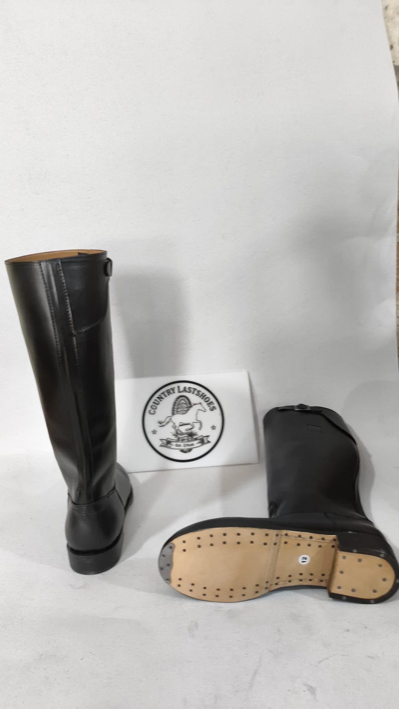 K&ouml;nnte beinhalten: Schwarze Lederreitstiefel, einer aufrecht und einer liegend, mit heller Sohle. Die Stiefel haben einen hohen Schaft und eine runde Spitze. Ein wei&szlig;es Schild mit dem Text "COUNTRY LASTSHOES" ist sichtbar.