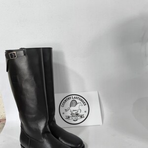 K&ouml;nnte beinhalten: Ein Paar hohe, schwarze Lederreitstiefel. Die Stiefel haben einen Schnallenverschluss oben und einen niedrigen Absatz. Eine wei&szlig;e Karte mit dem Text "COUNTRY LASTSHOES" und einem Logo ist sichtbar.
