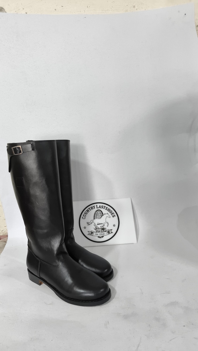 K&ouml;nnte beinhalten: Ein Paar hohe, schwarze Lederreitstiefel. Die Stiefel haben einen Schnallenverschluss oben und einen niedrigen Absatz. Eine wei&szlig;e Karte mit dem Text "COUNTRY LASTSHOES" und einem Logo ist sichtbar.