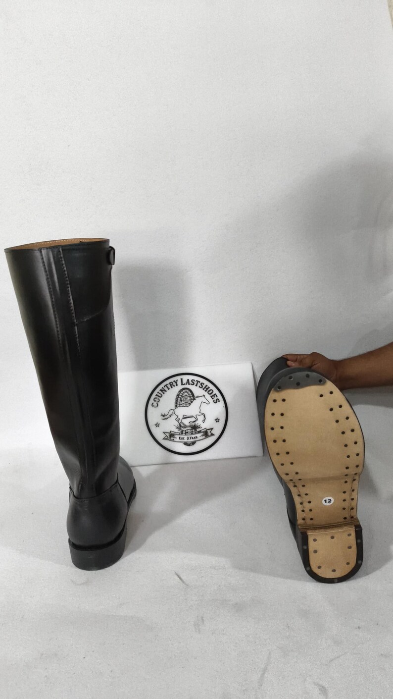 K&ouml;nnte beinhalten: Schwarze Lederreitstiefel mit gen&auml;hter Sohle. Die Stiefel sind hoch, reichen bis zum Knie und haben ein Schnallendetail. Die Sohle eines Stiefels ist sichtbar und zeigt die Gr&ouml;&szlig;e 12. Ein Schild mit dem Text "COUNTRY LASTSHOES" ist ebenfalls sichtbar.