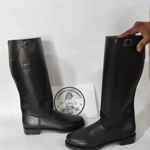 K&ouml;nnte beinhalten: Ein Paar schwarze Lederreitstiefel. Die Stiefel sind hoch, mit glatter Oberfl&auml;che und Schnallendetail oben. Eine wei&szlig;e Karte mit dem Text "COUNTRY LASTSHOES" und einem Pferdelogo ist sichtbar.