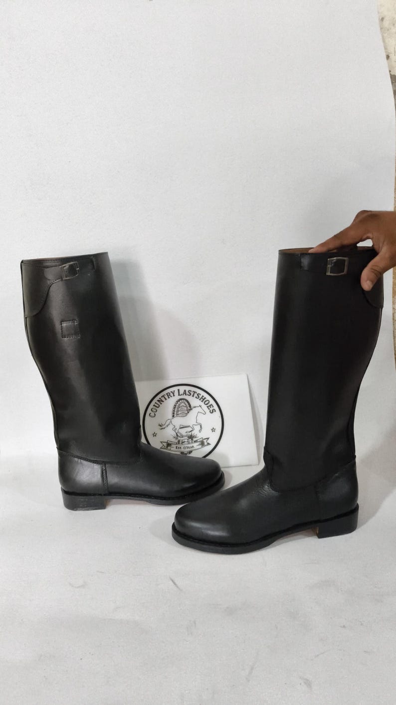 K&ouml;nnte beinhalten: Ein Paar schwarze Lederreitstiefel. Die Stiefel sind hoch, mit glatter Oberfl&auml;che und Schnallendetail oben. Eine wei&szlig;e Karte mit dem Text "COUNTRY LASTSHOES" und einem Pferdelogo ist sichtbar.