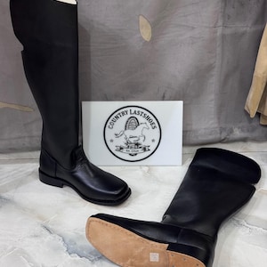 Puede incluir: Un par de botas altas de cuero negro con punta cuadrada y tacón plano. Una bota está de pie mientras que la otra está tumbada de lado, mostrando una suela de cuero color canela. Un letrero rectangular blanco muestra el texto "COUNTRY LASTSHOES".