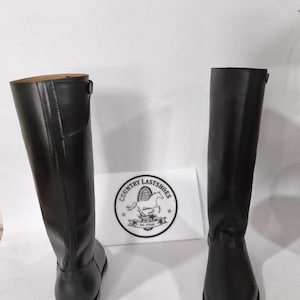 K&ouml;nnte beinhalten: Zwei schwarze Lederreitstiefel. Die Stiefel sind hoch, mit glatter Oberfl&auml;che und leichtem Absatz. Die Stiefel stehen auf einer wei&szlig;en Oberfl&auml;che. Ein wei&szlig;es Schild mit dem Text "COUNTRY LASTSHOES" befindet sich zwischen den Stiefeln.