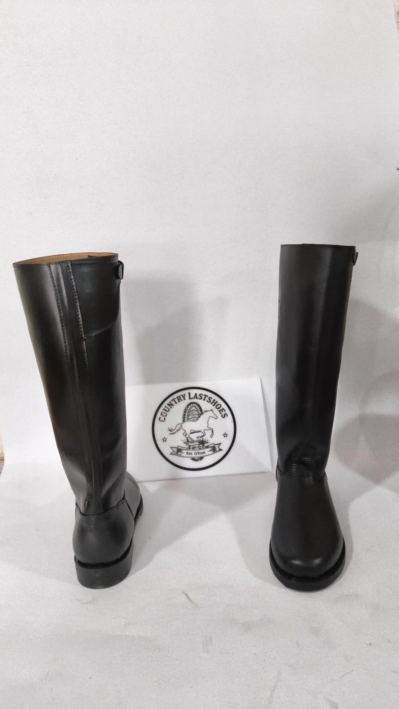 K&ouml;nnte beinhalten: Zwei schwarze Lederreitstiefel. Die Stiefel sind hoch, mit glatter Oberfl&auml;che und leichtem Absatz. Die Stiefel stehen auf einer wei&szlig;en Oberfl&auml;che. Ein wei&szlig;es Schild mit dem Text "COUNTRY LASTSHOES" befindet sich zwischen den Stiefeln.