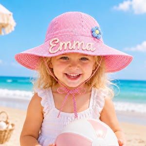 May include: A pink and white pinstriped sun hat featuring the name Emma in white script lettering on the front, a small blue flower accent on the side, and a matching pinstriped chin strap tied in a bow under the chin for a secure fit.