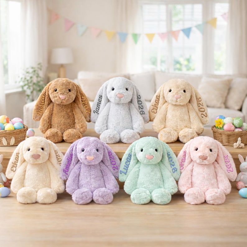 May include: A collection of plush bunny rabbits in various pastel colors, including brown, white, yellow, lavender, mint green, and pink. Each bunny has a name and the year 2026 embroidered on its ear. The bunnies are arranged on a wooden surface.
