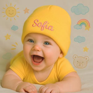 May include: A smiling baby wearing a yellow beanie embroidered with the name "Sofia" in pink. The baby is also wearing a yellow onesie. The background features cartoon illustrations of a sun, stars, clouds, a rainbow, and a bear.