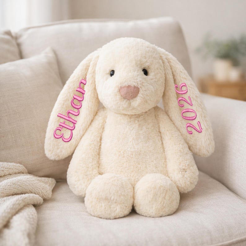 May include: A cream-colored plush bunny rabbit with long floppy ears. The left ear has the name "Ethan" embroidered in pink, and the right ear has "2026" embroidered in pink. The bunny is sitting on a cream-colored chair.
