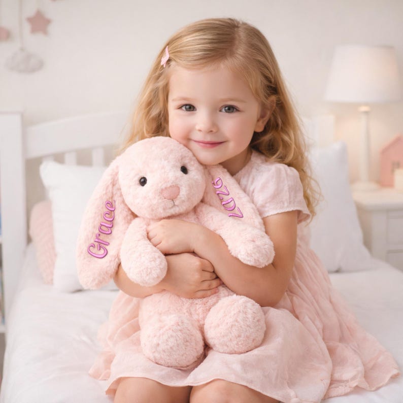 May include: A soft, pink plush bunny with the name "Grace" embroidered on one ear and the letters "9ZD7" on the other. The bunny is being held by a young child wearing a pink dress. The background includes a bed and a lamp.