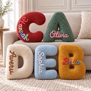 Può includere: Cinque cuscini in peluche dalla consistenza testurizzata a forma di lettera, nei colori rosso, verde, crema, azzurro e giallo senape. La C rossa riporta il nome Hannah, la A verde Olivia, la D crema Avery, la E blu Sophia e la B gialla Bella.