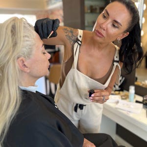 May include: A makeup artist applying makeup to a client in a salon setting. The artist, wearing a black glove, is focused on the client's eyebrow. The client is wearing a black cape. The artist is wearing a white jumpsuit.