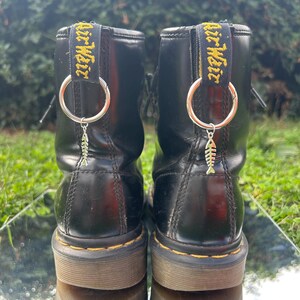 May include: Pair of black leather ankle boots with yellow stitching and pull tabs. Each boot has a silver ring and a small fish charm. The boots are reflected on a mirrored surface.