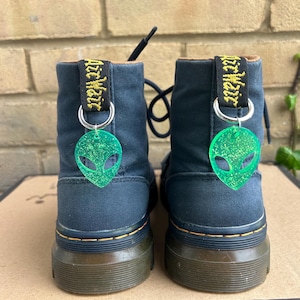 May include: A pair of navy blue ankle boots with yellow stitching and a thick sole. Each boot has a green glitter alien head charm hanging from the back. The words "Air Wair" are printed on the back of the boots.