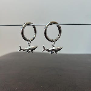 May include: A pair of silver-colored hoop earrings. Each earring features a small, detailed shark charm dangling from the hoop. The sharks are facing the same direction, with fins and tail visible.