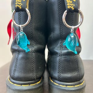 May include: Pair of black leather boots with yellow stitching and a black and yellow pull tab. Each boot has a silver ring with a blue dolphin charm attached. Red laces are visible.