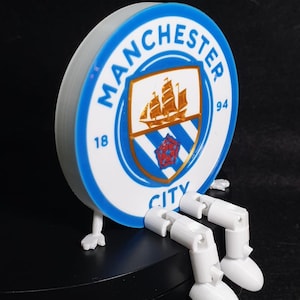 Op de afbeelding: Een 3D-geprint Manchester City-logo met witte benen en voeten. Het cirkelvormige logo heeft een blauwe rand met "MANCHESTER CITY" in het wit. Het midden toont een schip en de nummers "18" en "94".