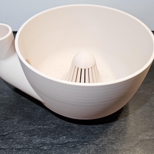May include: A white, 3D-printed kitchen tool with a bowl-like structure, a central strainer, and a curved spout. The design is modern and functional, suitable for food preparation. The object is made of a light-coloured plastic.