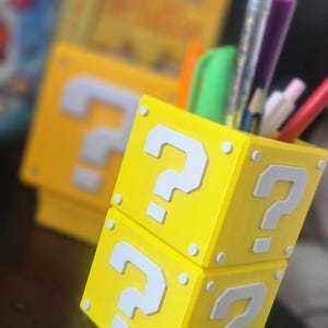 Puede incluir: Un portalápices amarillo con forma de bloque de signo de interrogación de la serie de videojuegos Super Mario. El bloque tiene signos de interrogación blancos y detalles blancos. El portalápices está lleno de bolígrafos, lápices y rotuladores.