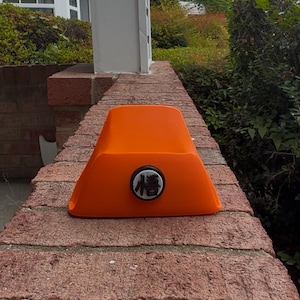 Peut inclure: Objet orange vif, de forme trapézoïdale, avec un emblème circulaire noir et blanc représentant un caractère japonais. L'objet repose sur un mur de briques rouges, avec de la verdure en arrière-plan.