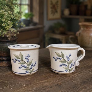 May include: A ceramic sugar bowl with a lid and a matching creamer pitcher, both featuring a speckled off-white finish, a painted blue berry and green leaf botanical pattern, and a thin blue horizontal line near the base of each piece.
