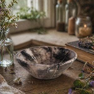 May include: A rustic, handcrafted bowl with a marbled gray and black pattern. The bowl sits on a weathered wooden surface, surrounded by dried flowers, a glass vase, and a vintage book. The bowl's diameter is approximately 8 inches.