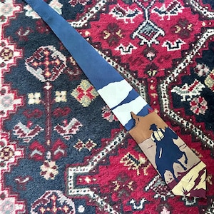 May include: A necktie featuring a desert landscape scene with a silhouette of a person riding a horse, a brown rock formation, and a blue sky. The upper portion of the tie is a solid dark blue with a subtle tonal pattern, tapering to a pointed end.