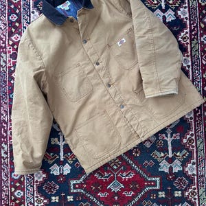 May include: A tan work jacket with a black collar and multiple pockets. The jacket has a button-up front and a small logo patch on the chest. The jacket is made of a durable material.